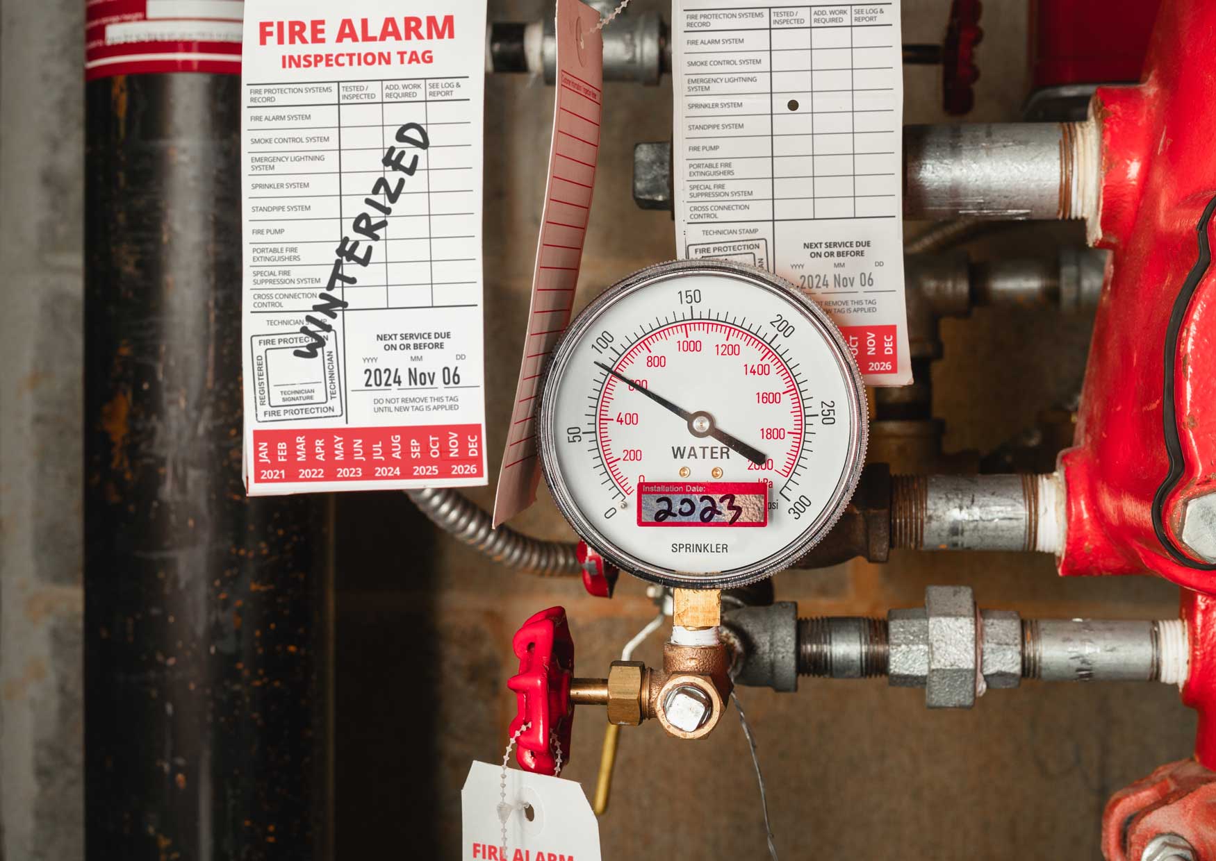 What Is ‘Escape of Water’ and 7 Ways to Prevent Water Damage | Wint Blog: Building fire sprinkler system with inspection tags in water meter room. Close up of water gauge and pipes. Defocused Winterized label for parkade fire sprinklers system protection. Selective focus.